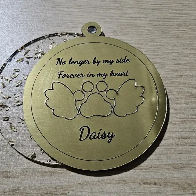 Round pet memorial ornament cut from brushed gold two-tone acrylic over a clear gold foil flake layer, engraved with a winged paw, No longer by my side, Forever in my heart, and the name Daisy