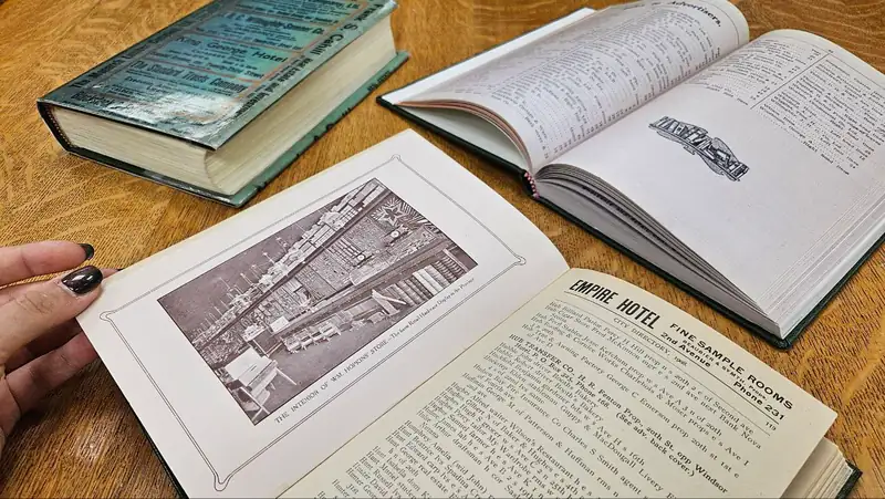 A hand opening an early twentieth century city directory beside two other open volumes with printed store illustrations
