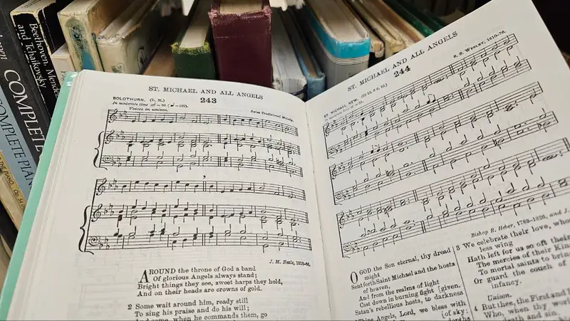 An open hymnal showing engraved musical notation and verse, with music books on the shelf behind